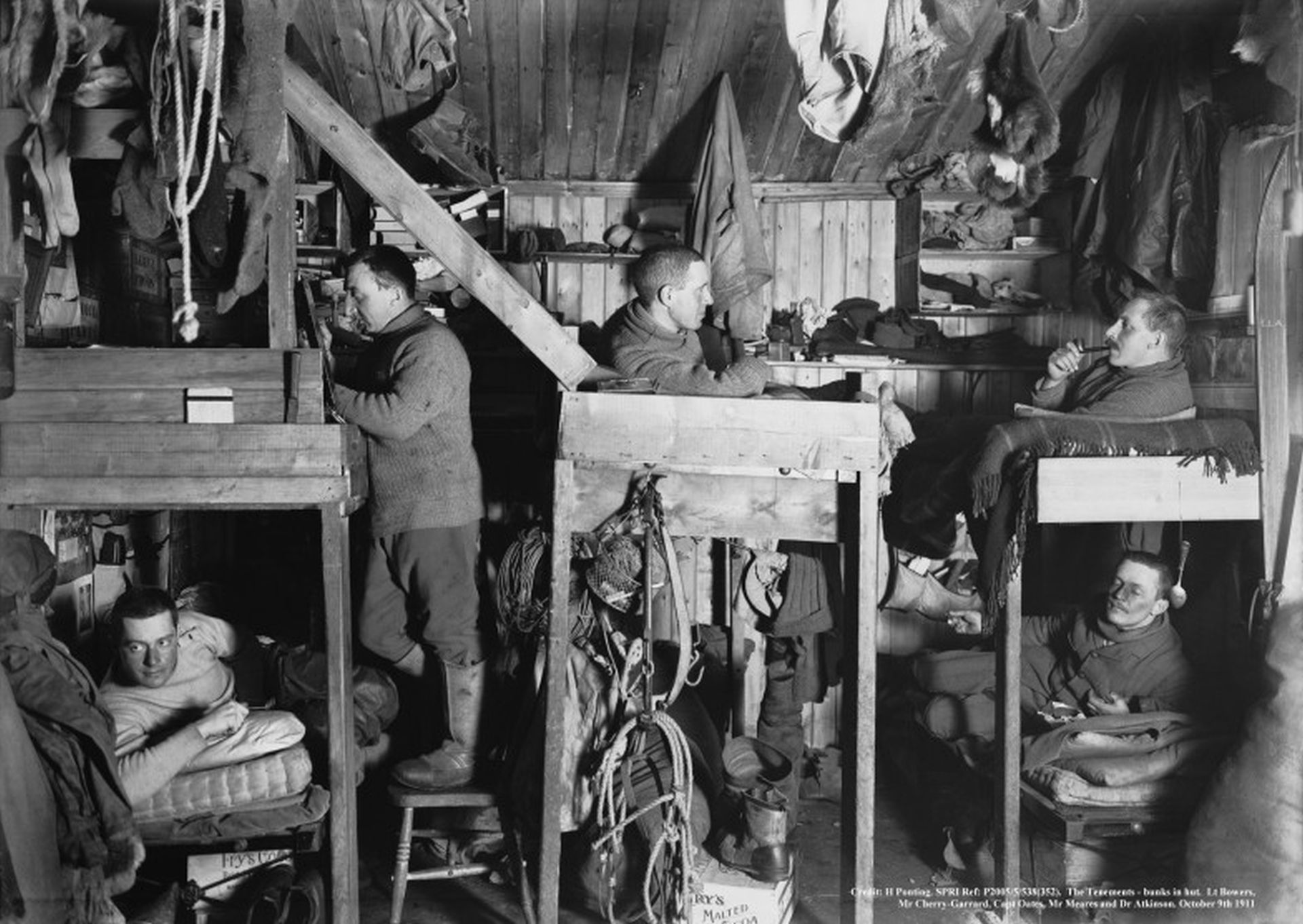 In the interior of the hut, Cherry-Garrard, Oates, Meares and Atkinson lie on bunks, Bowers stands on a chair next to his.