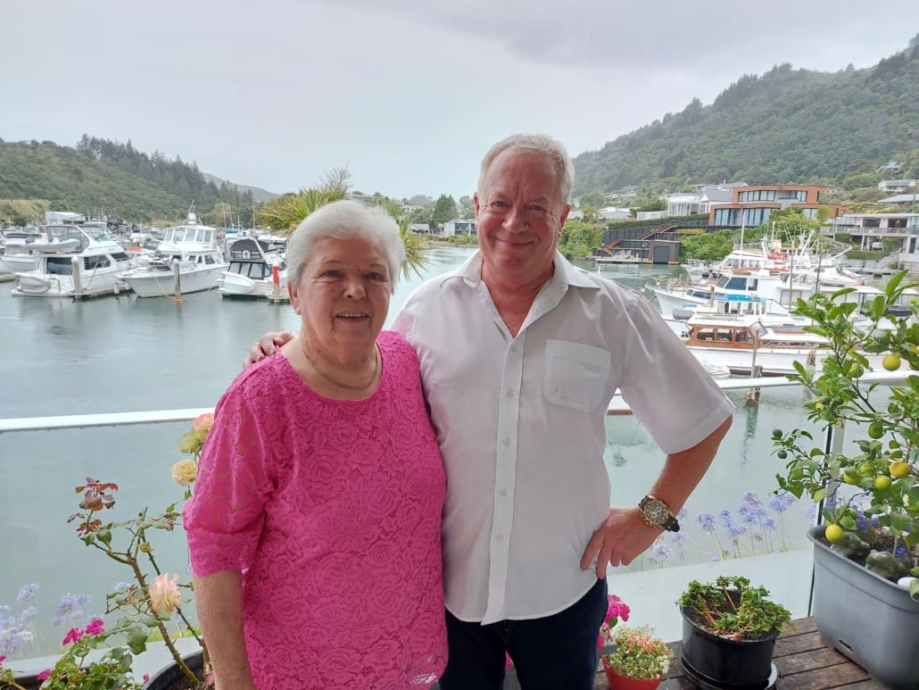 A couple standing on a balcony, with a marina and homes in the background.