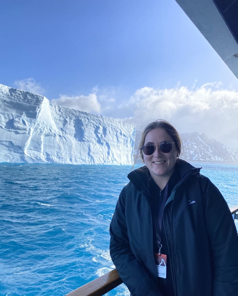 A woman standing on the balcony of a boat, with a large iceberg in the background.
