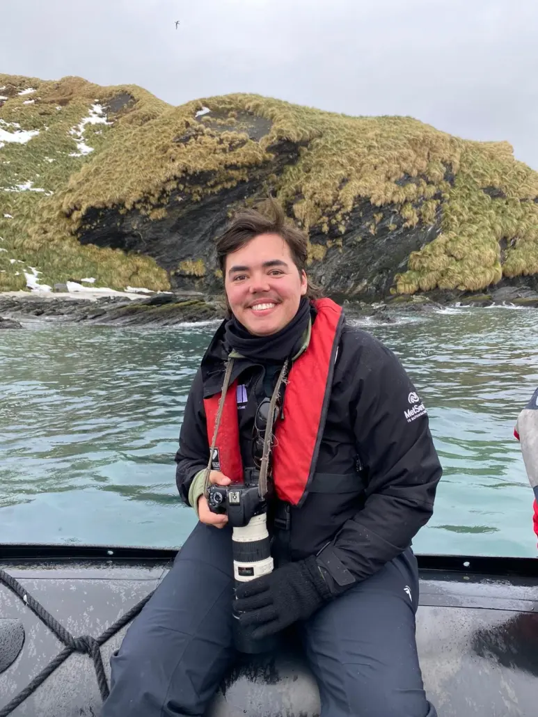 A person sits on the edge of a boat, holding a camera, with water and land behind them.