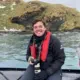 A person sits on the edge of a boat, holding a camera, with water and land behind them.