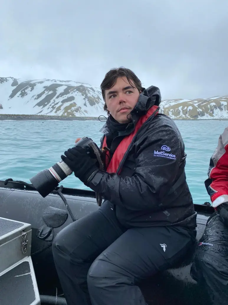 A person sits in a boat, holding a camera, looking at something out of frame, with snow covered mountains in the background.