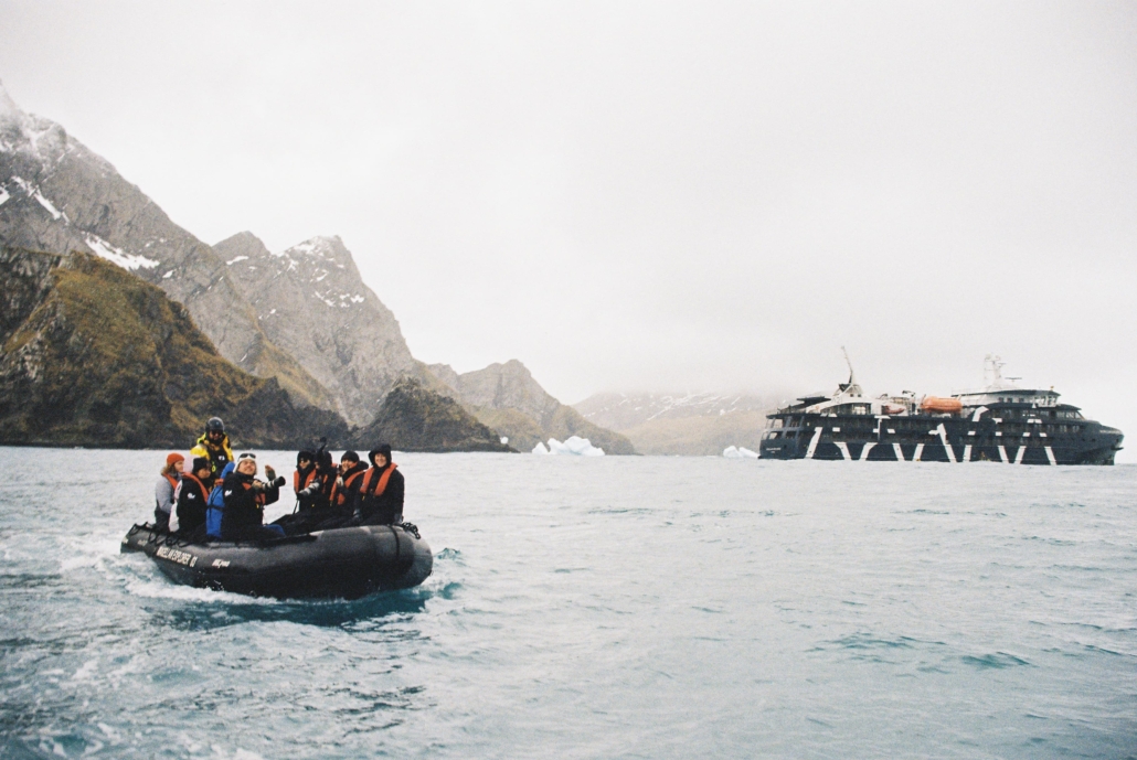 A zodiac with passengers. Mountains and a large cruise ship in the background.